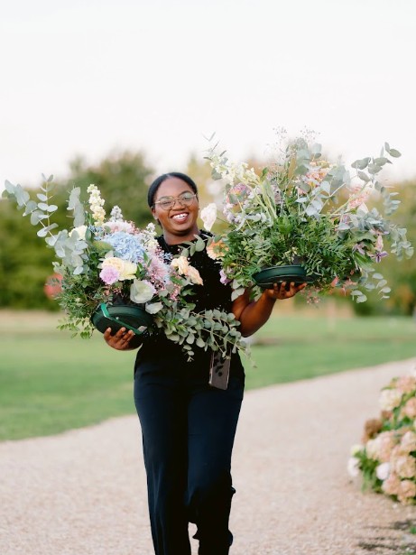 Armande souriante, portant deux grandes compositions florales dans un jardin