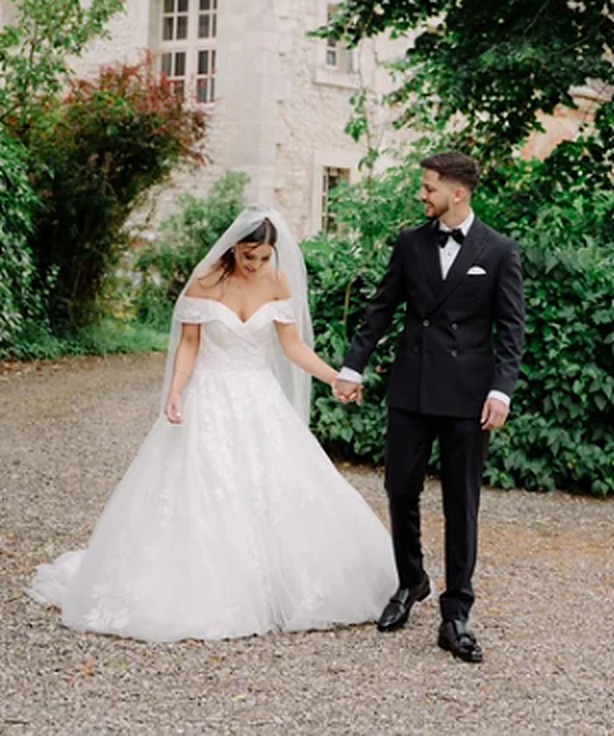 Aline and Denis hand in hand on a château driveway, wedding dress with embroidered flowers and black tuxedo