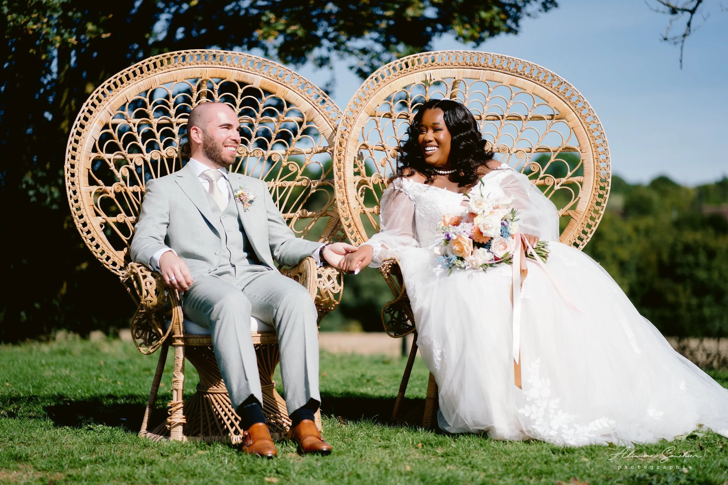 Newlyweds seated side by side on rattan armchairs, holding hands, outdoors