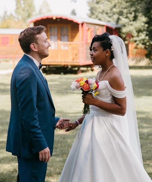 Camille and Patrick facing each other in a park, colourful bouquet, wooden caravans behind them